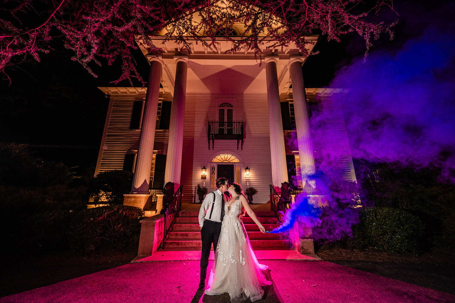 bride and groom kissing as they are leaving a wedding lit up by red and purple lights