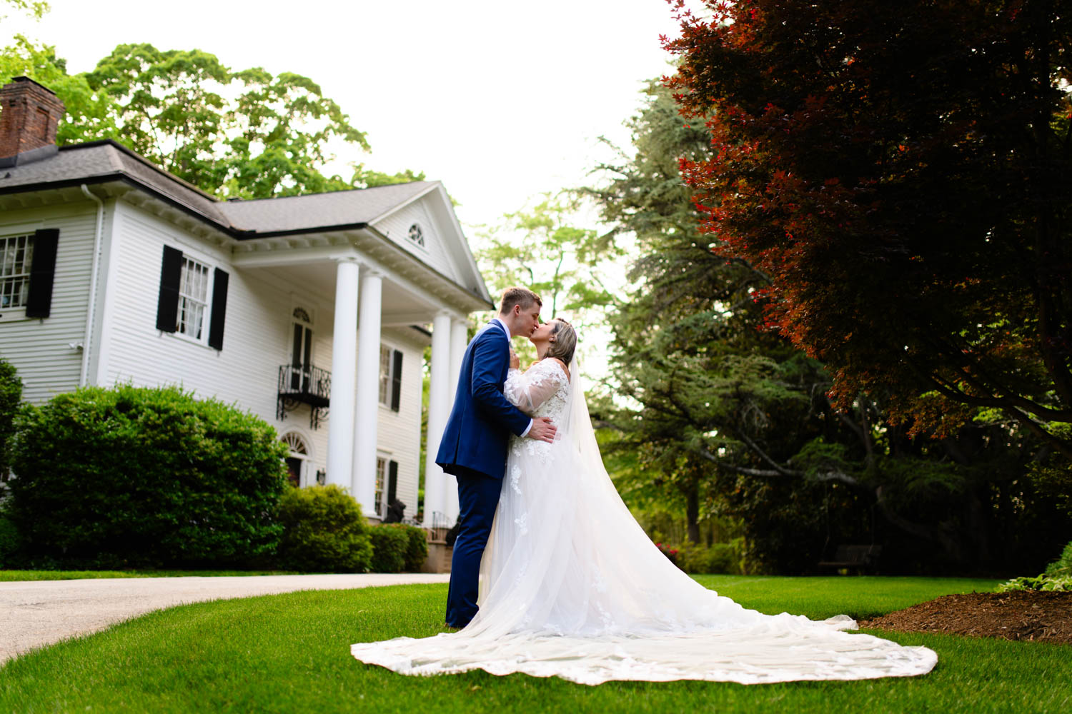 Couple in wedding clothes kissing with large white house in the background