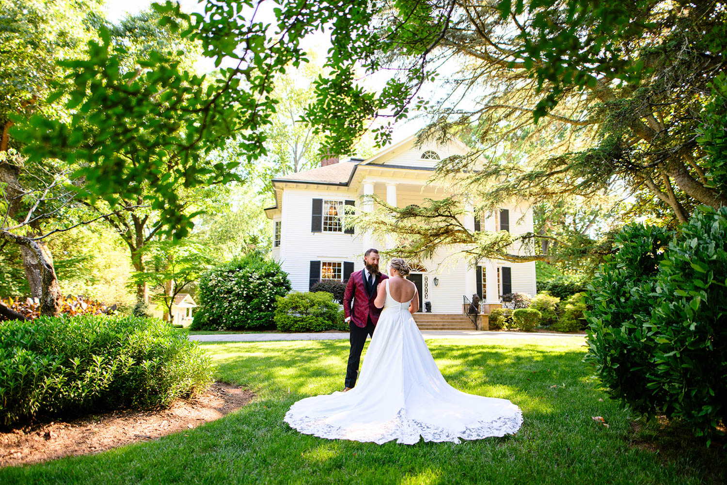 groom in red jacket and woman in wedding dress in front of white large house