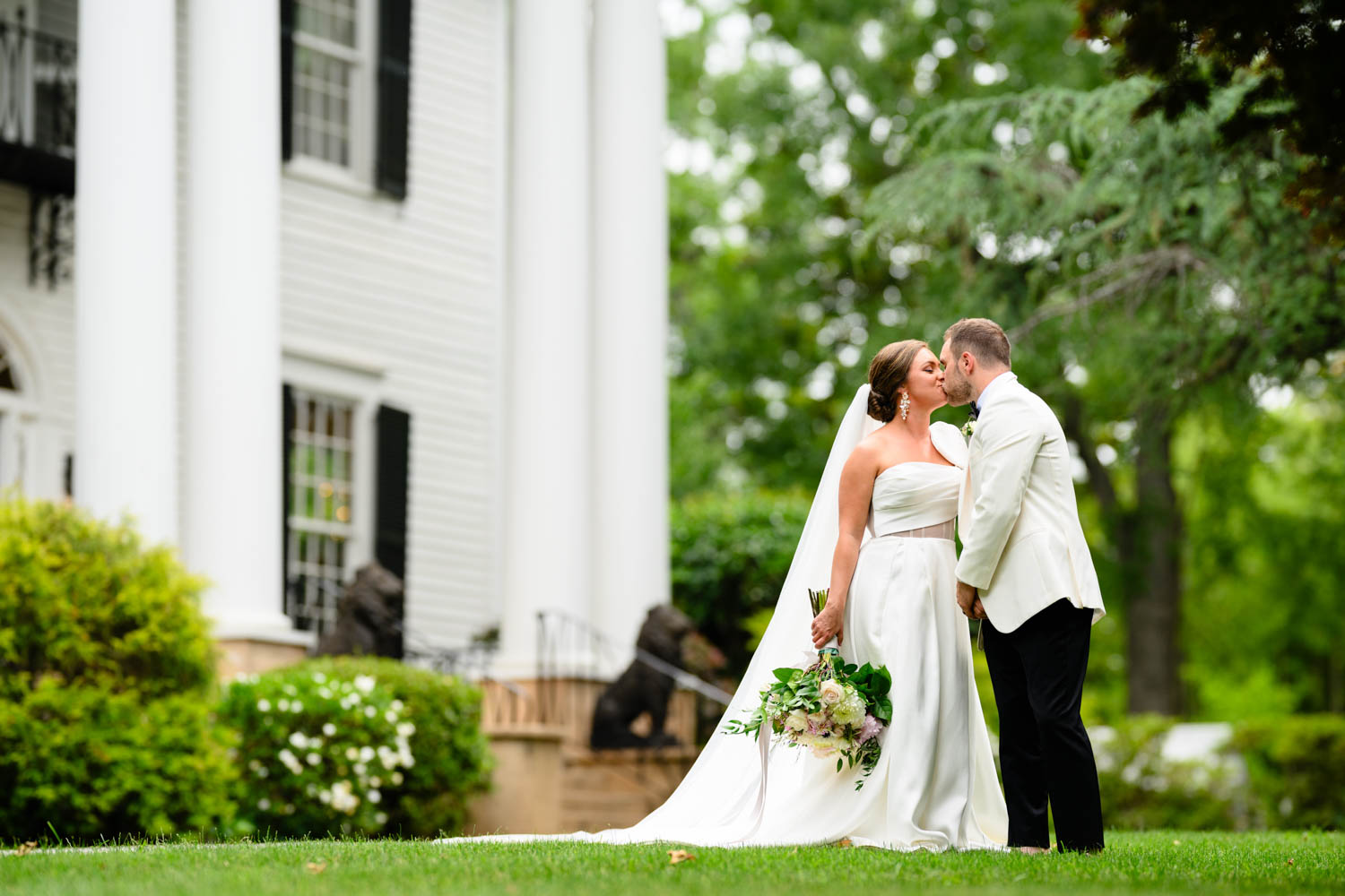 Couple kissing in wedding attire in front of a large white mansion